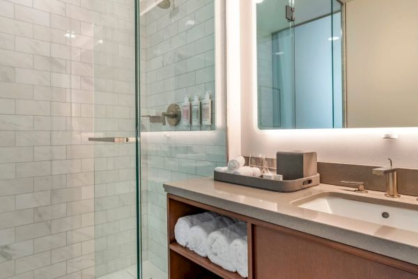 The image shows a modern bathroom with a glass-enclosed shower, a sink with a lighted mirror, and neatly arranged toiletries and towels.