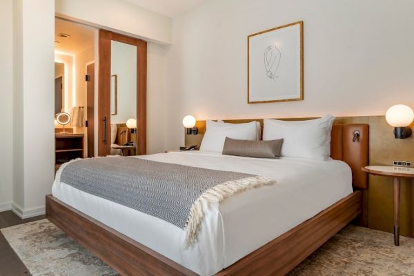 A modern bedroom with a neatly made bed, two bedside tables with lamps, a large mirror, and wall art above the headboard. Background shows a bathroom.