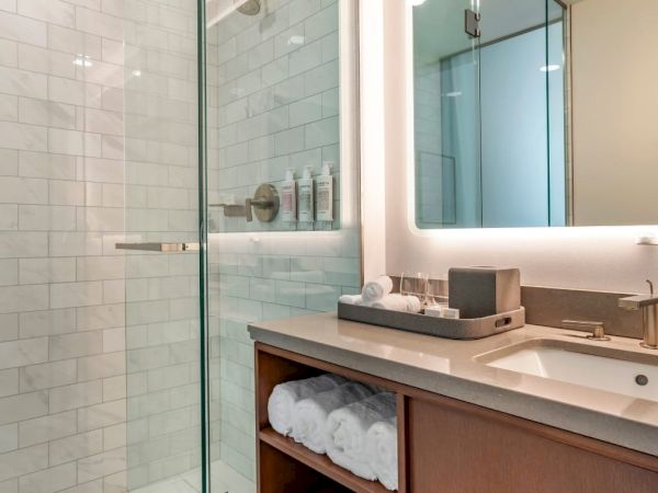 The image shows a modern bathroom with a glass-enclosed shower, a sink with a lighted mirror, and neatly arranged toiletries and towels.
