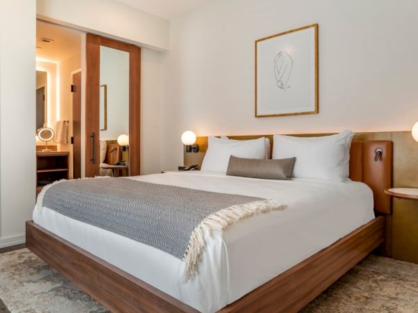 A modern bedroom with a neatly made bed, two bedside tables with lamps, a large mirror, and wall art above the headboard. Background shows a bathroom.