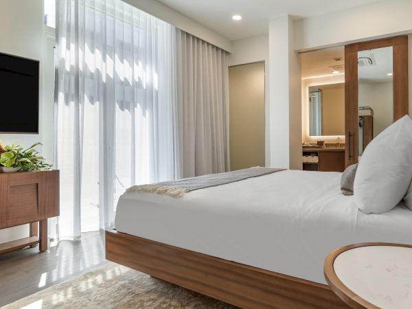 A modern bedroom featuring a large bed with white linens, a wooden nightstand with a plant, and a TV mounted on the wall.