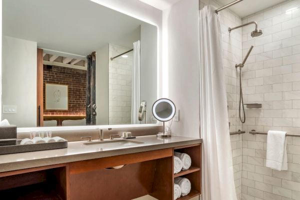 This image shows a modern bathroom with a large mirror, a vanity with a sink, shelves with towels, and a walk-in shower with white tiles and a curtain.