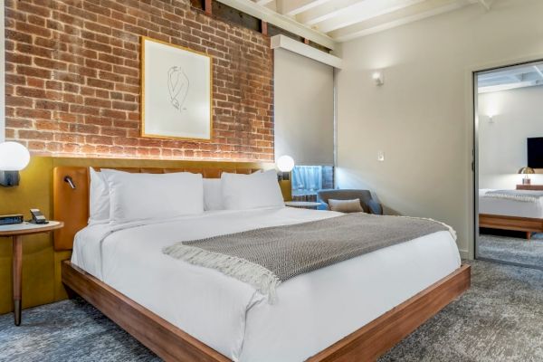 A modern bedroom features a large bed, brick accent wall, framed art, and a connecting room with another bed and TV visible through the open door.