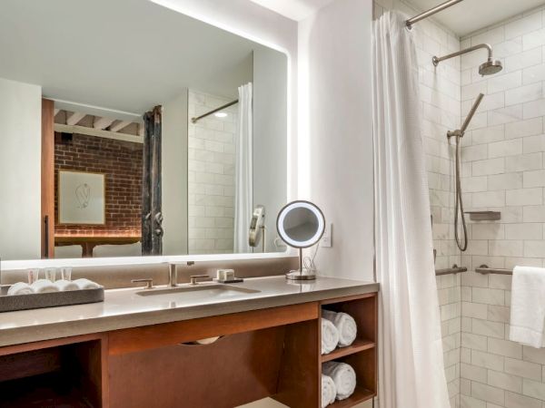 This image shows a modern bathroom with a large mirror, a vanity with a sink, shelves with towels, and a walk-in shower with white tiles and a curtain.