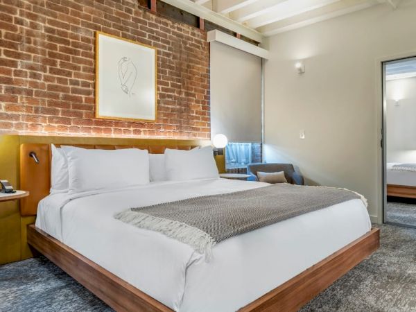 A modern bedroom features a large bed, brick accent wall, framed art, and a connecting room with another bed and TV visible through the open door.