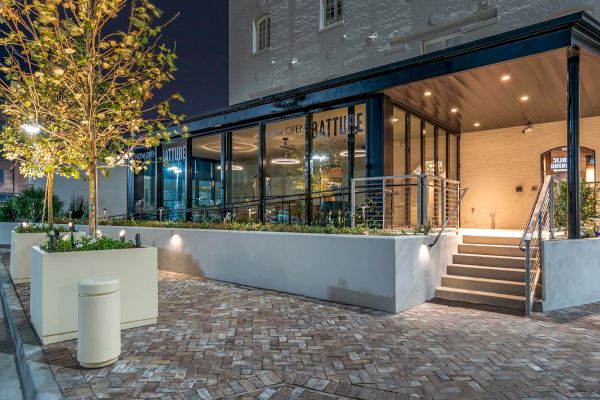 The image shows a modern cafe exterior at night with large glass windows, outdoor seating, potted plants, and a staircase leading to the entrance.