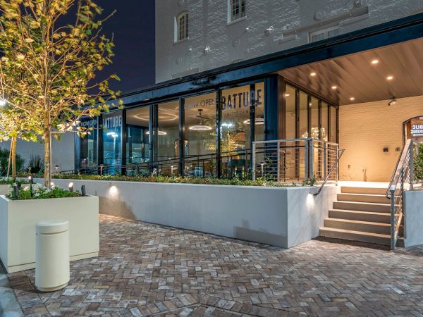 The image shows a modern cafe exterior at night with large glass windows, outdoor seating, potted plants, and a staircase leading to the entrance.