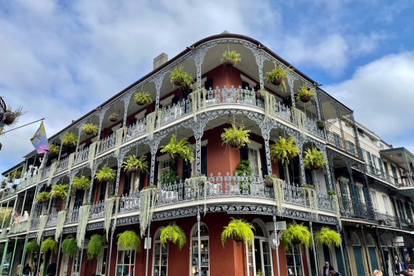 A historic building features ornate ironwork and balconies adorned with hanging plants, set against a partly cloudy blue sky.