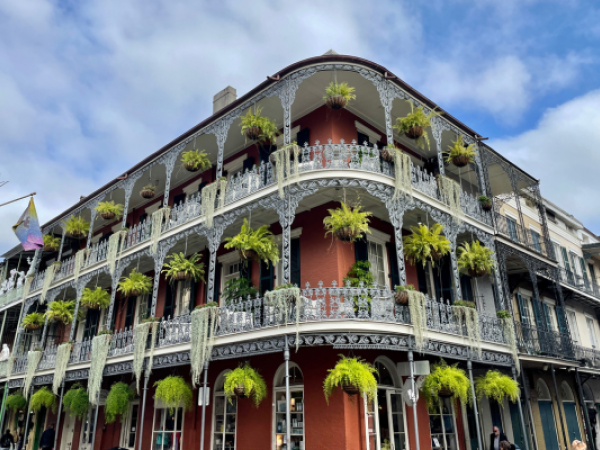 A historic building features ornate ironwork and balconies adorned with hanging plants, set against a partly cloudy blue sky.