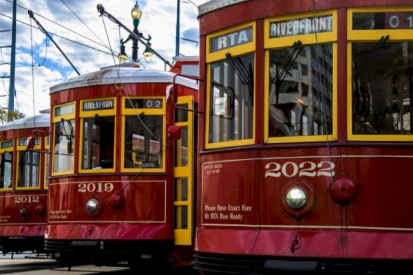 The image shows three red streetcars with yellow accents on a city street under a blue sky with clouds.