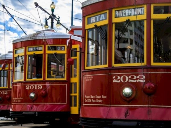 The image shows three red streetcars with yellow accents on a city street under a blue sky with clouds.