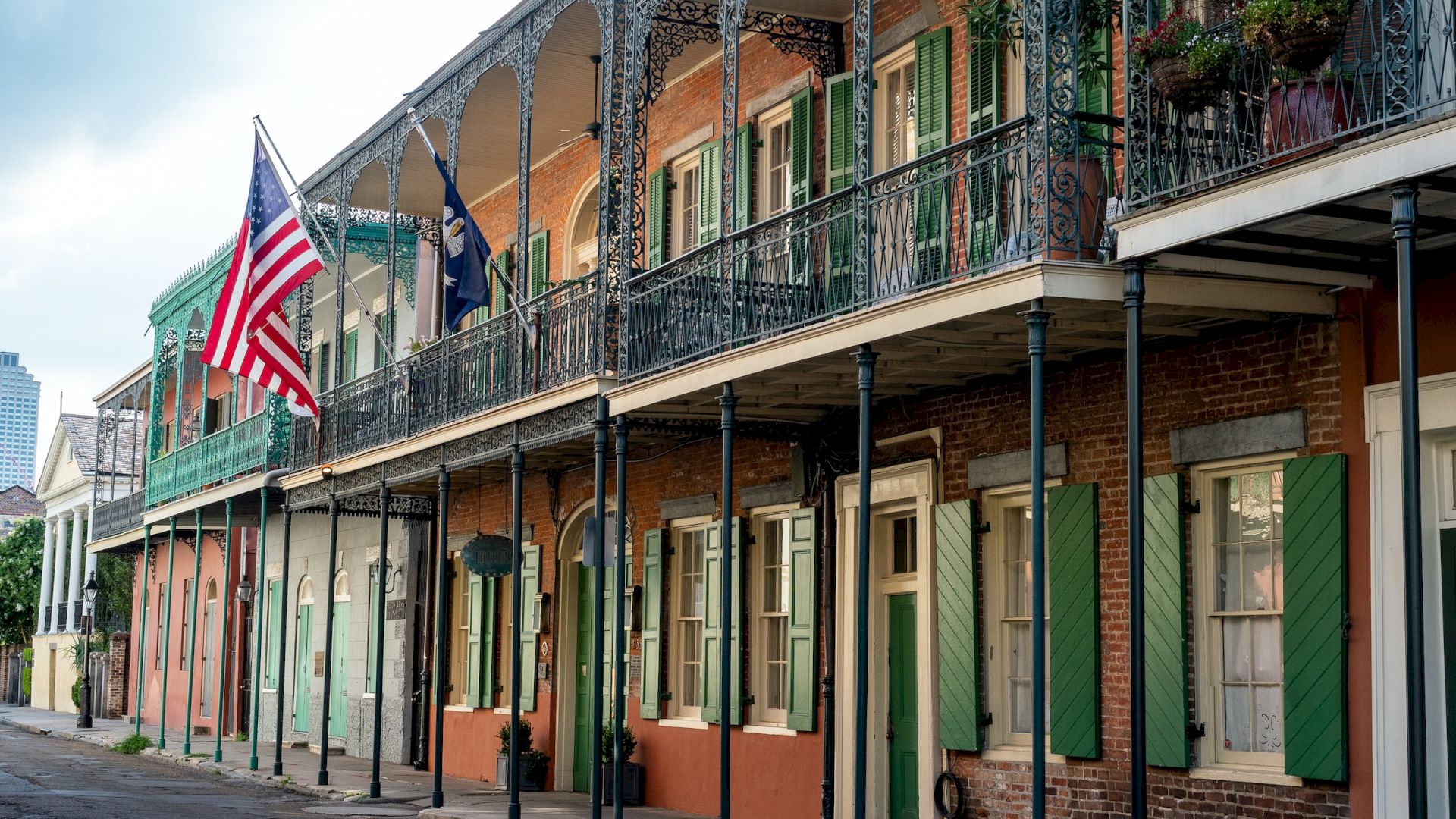 A street with historic, colorful, French-style buildings and balconies, featuring the American flag, green shutters, and intricate ironwork.