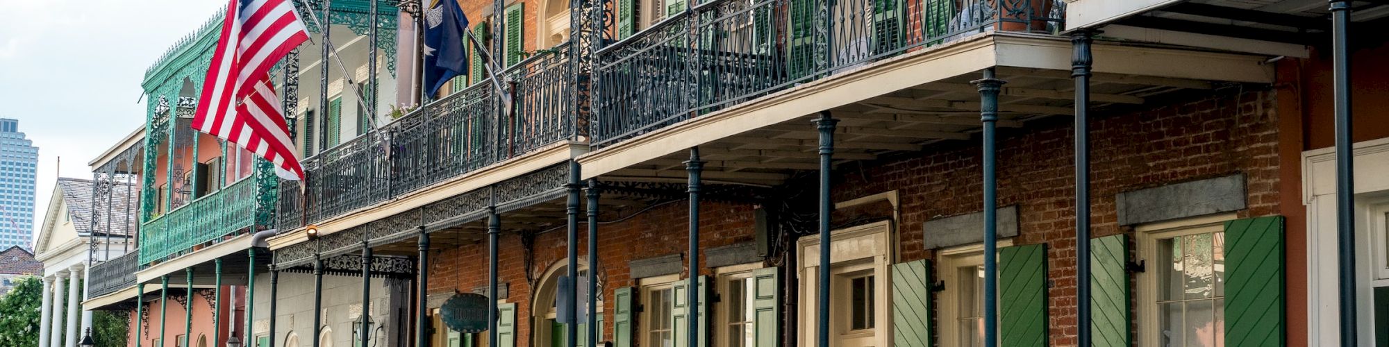 A street with historic, colorful, French-style buildings and balconies, featuring the American flag, green shutters, and intricate ironwork.