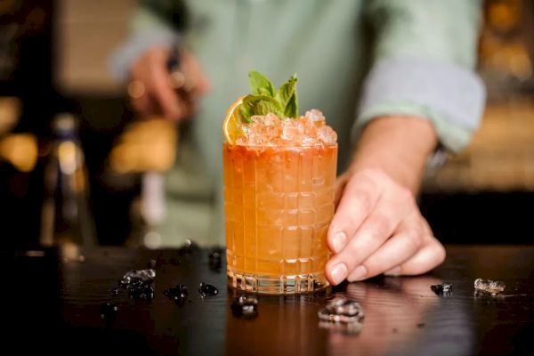 A person is serving a cocktail garnished with mint leaves and an orange slice. The drink is in a glass on a bar counter with scattered ice cubes.