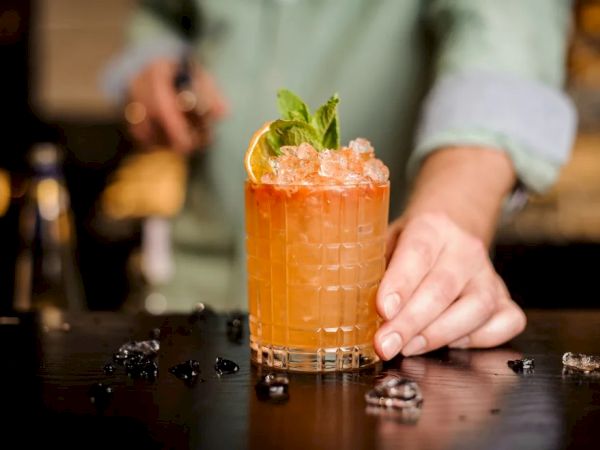 A person is serving a cocktail garnished with mint leaves and an orange slice. The drink is in a glass on a bar counter with scattered ice cubes.