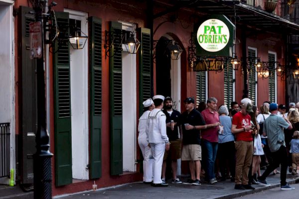 People are gathered outside a bar named Pat O'Brien's, situated in a building with green shutters. Some are in casual attire, others in uniforms.