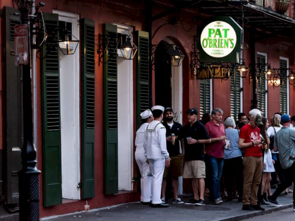 People are gathered outside a bar named Pat O'Brien's, situated in a building with green shutters. Some are in casual attire, others in uniforms.