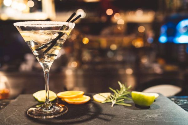 A cocktail in a martini glass with a salted rim and two straws, accompanied by citrus slices and herbs on a bar counter.