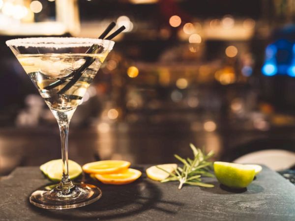 A cocktail in a martini glass with a salted rim and two straws, accompanied by citrus slices and herbs on a bar counter.
