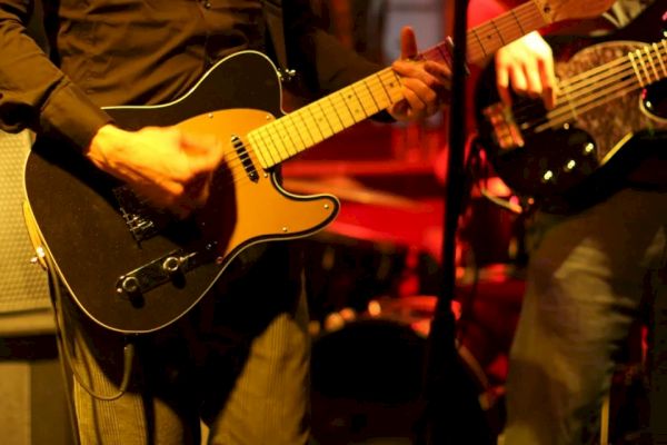 The image shows two musicians playing electric guitars on stage with a dimly lit background, suggesting a live performance.