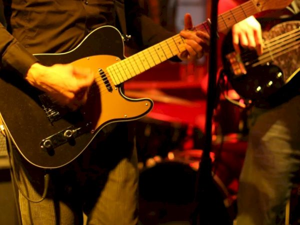 The image shows two musicians playing electric guitars on stage with a dimly lit background, suggesting a live performance.
