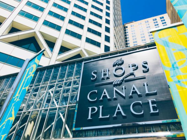 This image shows a sign reading "The Shops at Canal Place" with modern buildings in the background and colorful banners on either side.