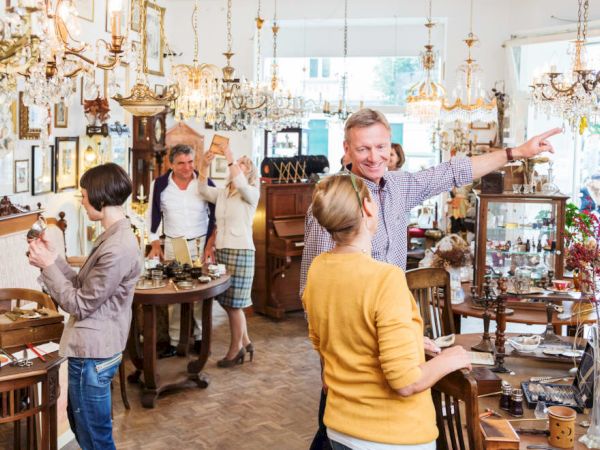 Several people browse and chat inside a well-lit antique shop filled with furniture, chandeliers, and various vintage items.