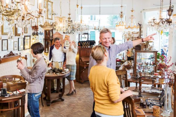 Several people browse and chat inside a well-lit antique shop filled with furniture, chandeliers, and various vintage items.