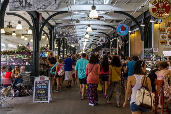 A bustling indoor market with numerous people walking and dining, various stalls selling goods, and signage hanging from the ceiling.