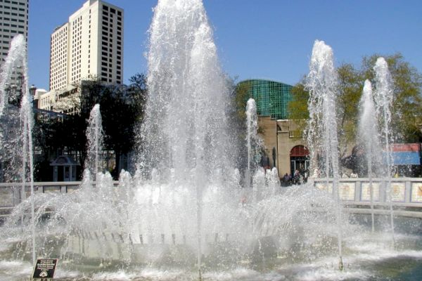 The image shows a large, ornate fountain with multiple water jets spraying upwards, set against a backdrop of tall buildings and trees.