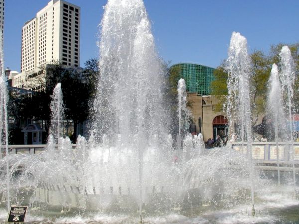 The image shows a large, ornate fountain with multiple water jets spraying upwards, set against a backdrop of tall buildings and trees.