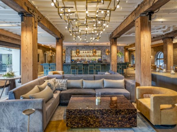 A modern lounge with wooden beams, a large sectional sofa, a unique coffee table, and a bar in the background, brightly lit by chandeliers.
