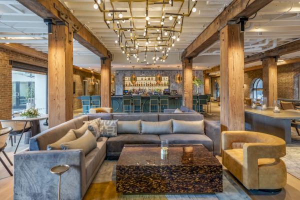 A modern lounge with wooden beams, a large sectional sofa, a unique coffee table, and a bar in the background, brightly lit by chandeliers.