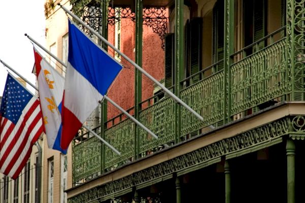 This image shows an American flag, a French flag, and another flag hanging from ornate iron balconies on a building.