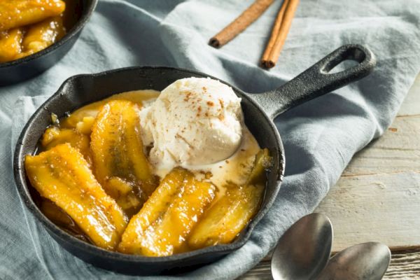 The image shows caramelized bananas topped with a scoop of vanilla ice cream in a small skillet, with cinnamon sticks and spoons nearby.