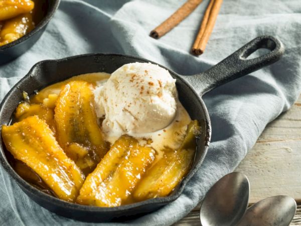 The image shows caramelized bananas topped with a scoop of vanilla ice cream in a small skillet, with cinnamon sticks and spoons nearby.