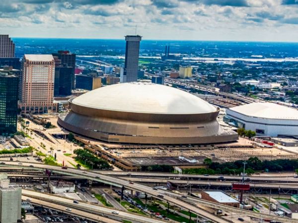 The image shows an aerial view of a modern city with a prominent dome-shaped stadium and surrounding buildings and highways under a partly cloudy sky.