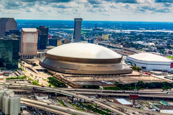 The image shows an aerial view of a modern city with a prominent dome-shaped stadium and surrounding buildings and highways under a partly cloudy sky.