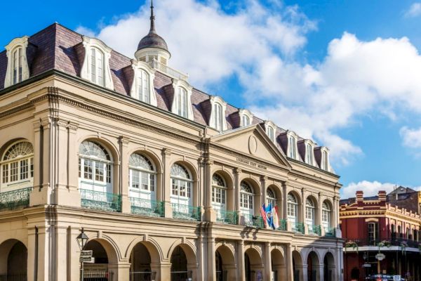 The image shows a historic building with arched windows and a dome, adorned with flags, set against a bright blue sky.