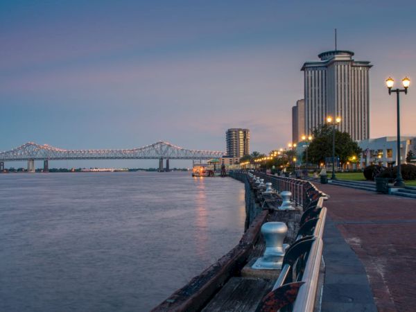 A waterfront view at dusk with a tall building, streetlights, a bridge in the background, and a walkway along the water.