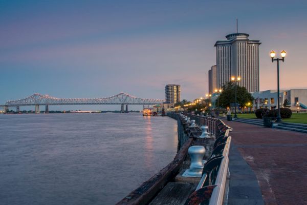 A waterfront view at dusk with a tall building, streetlights, a bridge in the background, and a walkway along the water.