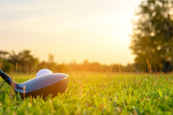 A golf ball on a tee with a golf club head nearby, set on a grassy field with trees and a bright sunset in the background.