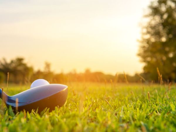 A golf ball on a tee with a golf club head nearby, set on a grassy field with trees and a bright sunset in the background.