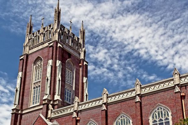 The image showcases a red-brick Gothic-style church with ornate windows and a tall tower against a blue sky with scattered clouds.