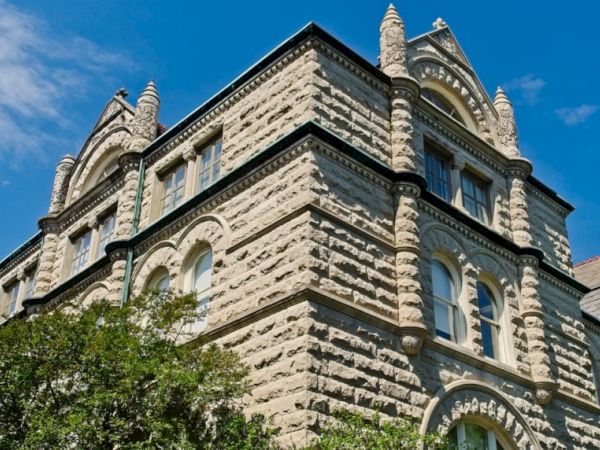 A stone building with Gothic architectural elements features arched windows and decorative spires, against a blue sky with a few scattered clouds.