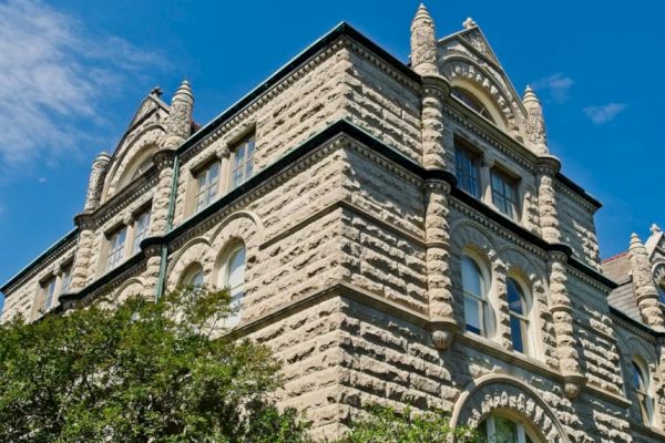 A stone building with Gothic architectural elements features arched windows and decorative spires, against a blue sky with a few scattered clouds.