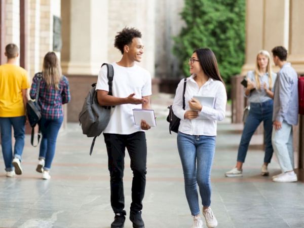 A group of students is walking and talking in an outdoor campus setting, with some holding notebooks and backpacks.