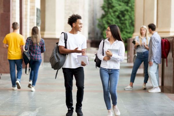 A group of students is walking and talking in an outdoor campus setting, with some holding notebooks and backpacks.