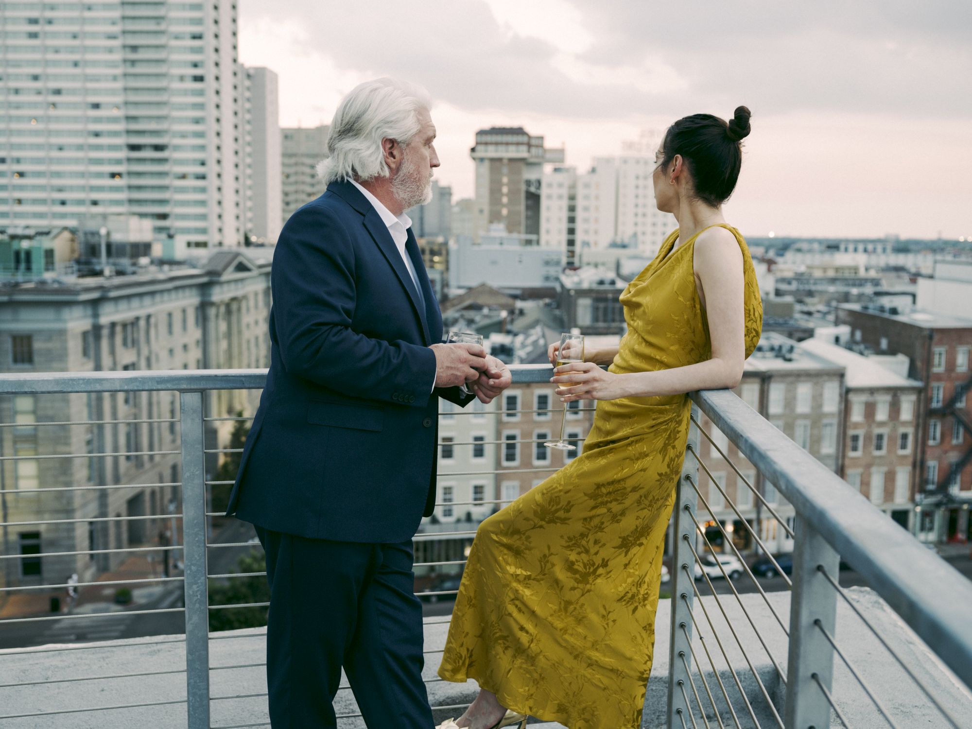 A man and woman in formal attire standing on a rooftop overlooking a city skyline, engaged in conversation with drinks in hand, during dusk.