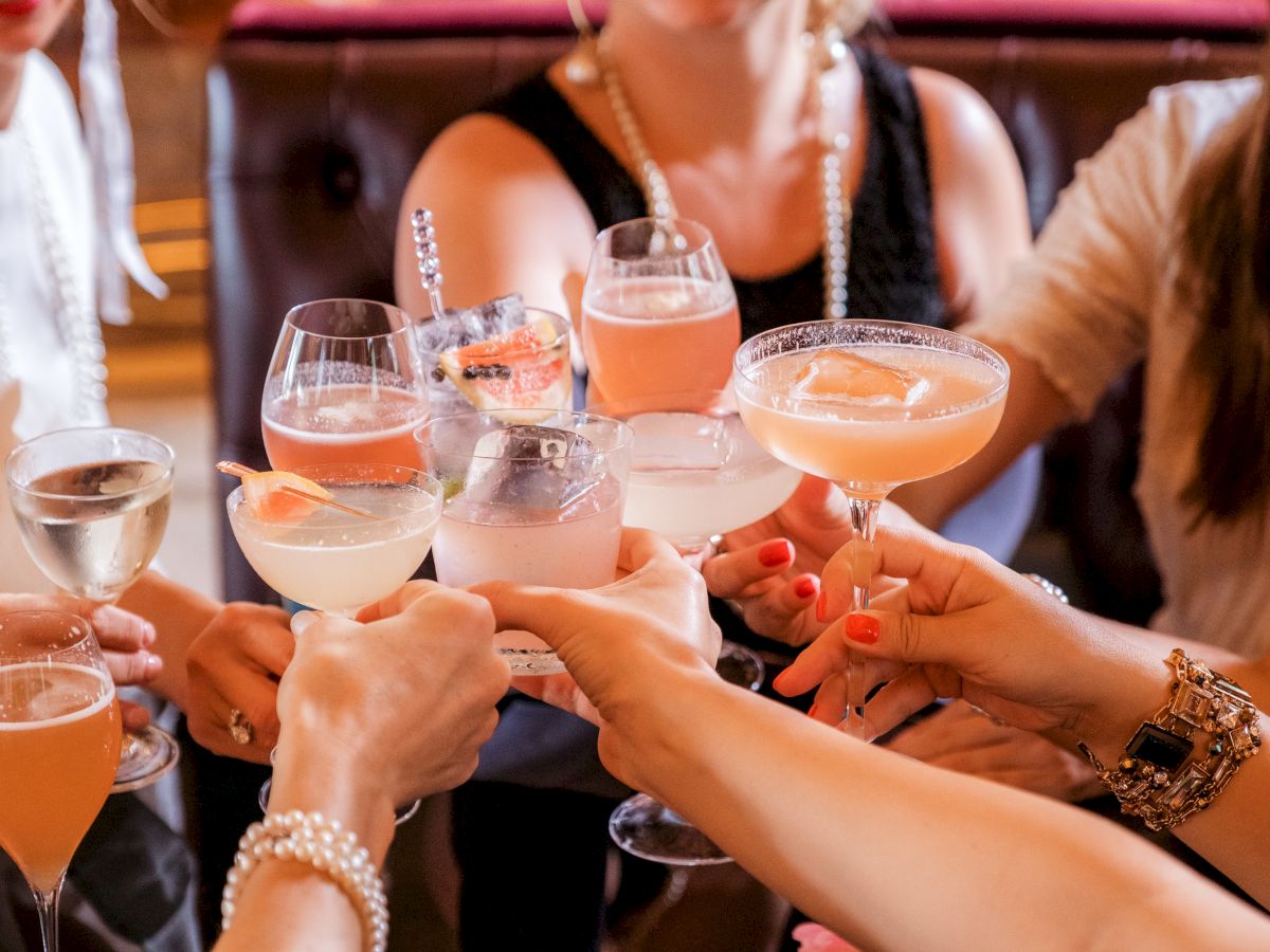 A group of people are toasting with various cocktails and glasses of wine in a celebratory setting, all hands raised, suggesting a gathering or celebration.
